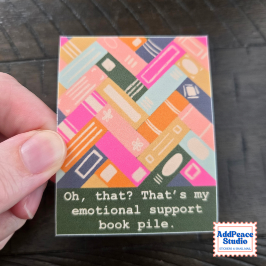 A hand holds a small square sticker featuring a colorful pattern of stacked books in pink, orange, teal, and green, with the text 'Oh, that? That's my emotional support book pile.' on a dark wood surface.