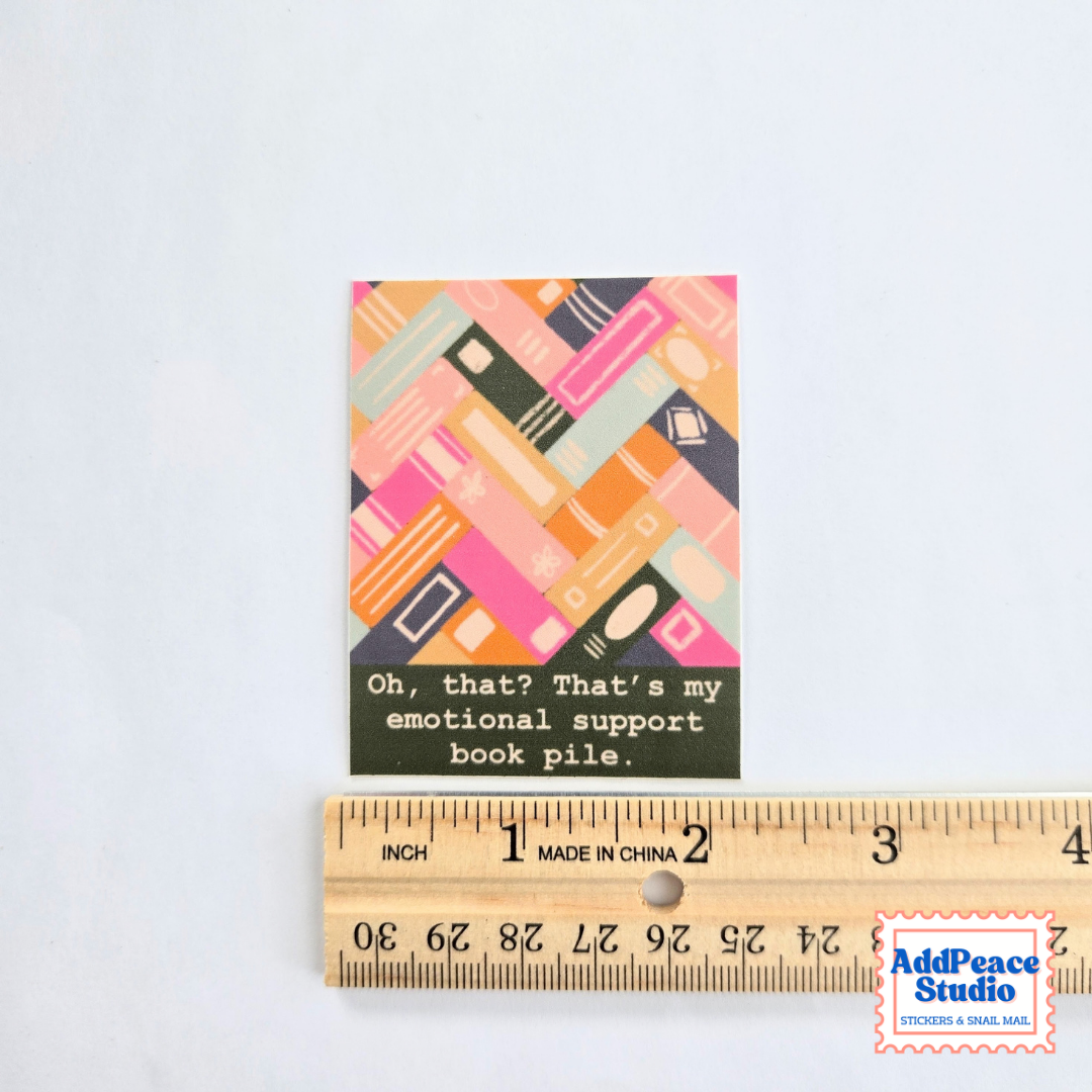 A small rectangular sticker with a colorful book pile illustration and the text 'Oh, that? That's my emotional support book pile.' is shown above a wooden ruler on a white background.
