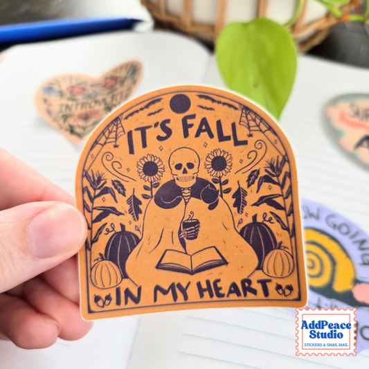 A hand holds a brown sticker with and illustration of a skeleton reading a book and holding a mug surrounded by fall things with the text It's fall in my heart.