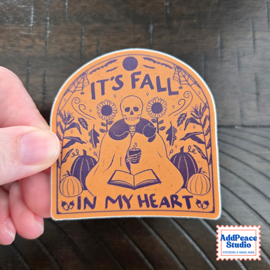 A hand holds a die-cut sticker with an autumn illustration. The sticker features a skeleton reading an open book, surrounded by pumpkins, sunflowers, and the text 'IT'S FALL IN MY HEART' in dark purple on an orange background.