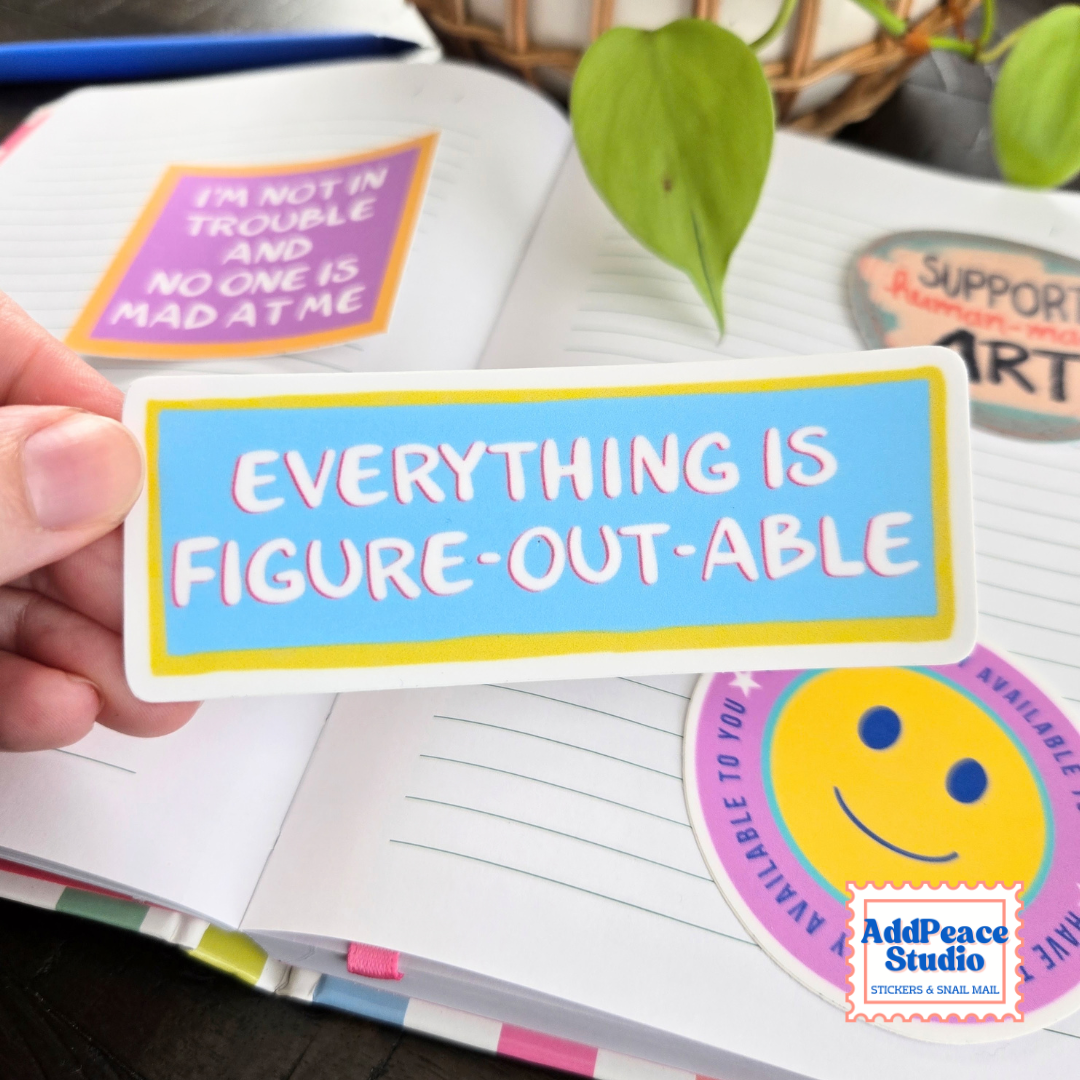 A hand holds a rectangular sticker with a light blue background, yellow border, and pink text reading 'EVERYTHING IS FIGURE-OUT-ABLE'. The scene shows an open notebook with colorful stickers, including a smiley face and supportive phrases, with a plant leaf in the background.