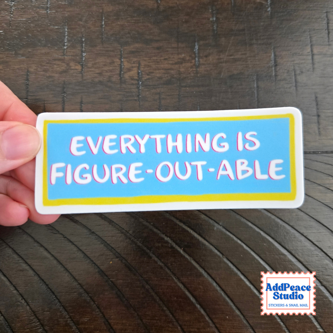 A hand holds a rectangular sticker with the phrase 'EVERYTHING IS FIGURE-OUT-ABLE' in pink letters on a light blue background with a yellow border, set against a dark wood surface.