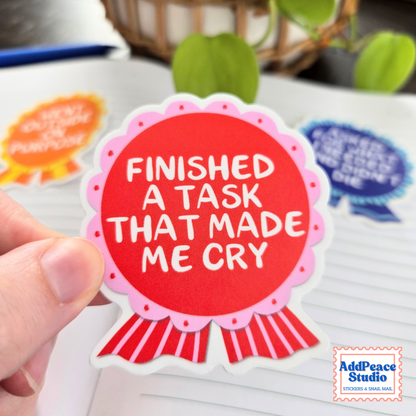 A hand holds a die-cut sticker shaped like a red ribbon badge with the text 'FINISHED A TASK THAT MADE ME CRY' in white. Other colorful stickers are visible in the blurred background on a white surface, with a plant and basket behind them.