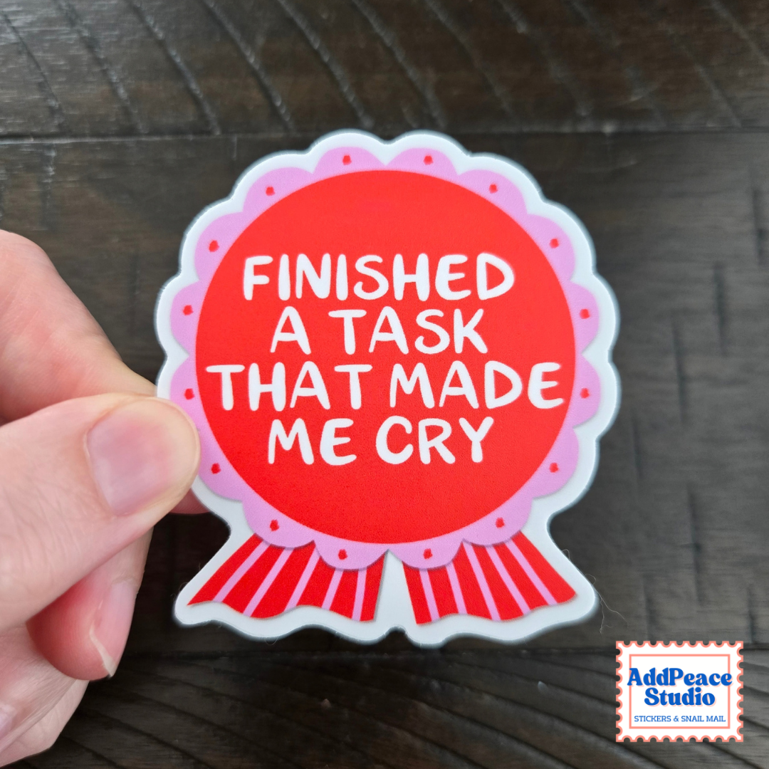 A hand holding a die-cut sticker with a red circular center, pink scalloped border, and white text reading 'FINISHED A TASK THAT MADE ME CRY' on a dark wood background. AddPeace Studio logo appears in the bottom right corner.