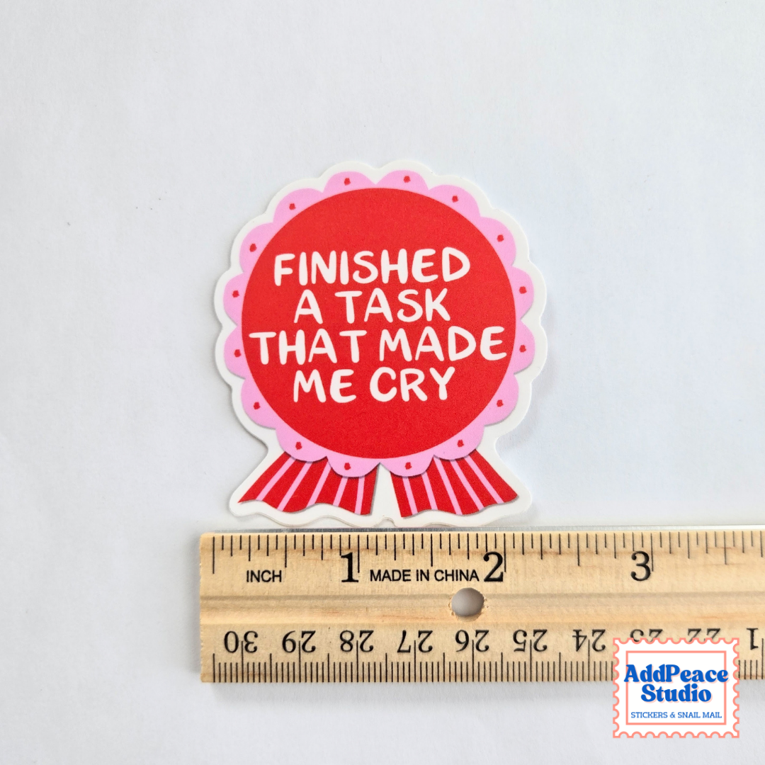A die-cut sticker with a red circular center, pink scalloped border, and red-and-white ribbon tail, printed with the phrase 'FINISHED A TASK THAT MADE ME CRY' in white text. The sticker is shown above a wooden ruler on a light gray background to indicate size.