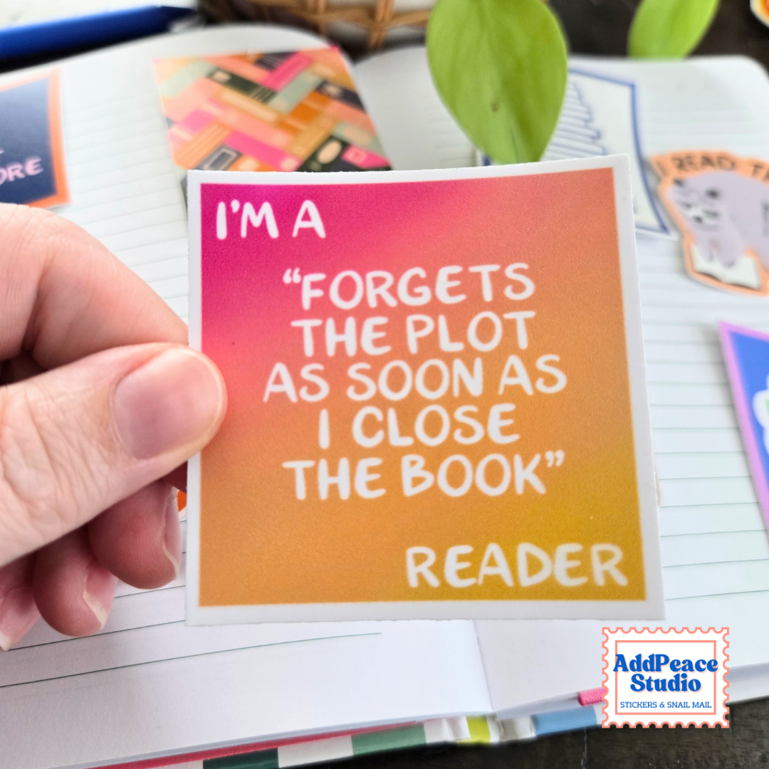 A hand holds a square sticker with a pink-to-orange gradient background and white text that reads, "I'M A 'FORGETS THE PLOT AS SOON AS I CLOSE THE BOOK' READER." The sticker is shown over an open notebook with other stationery items in the background.