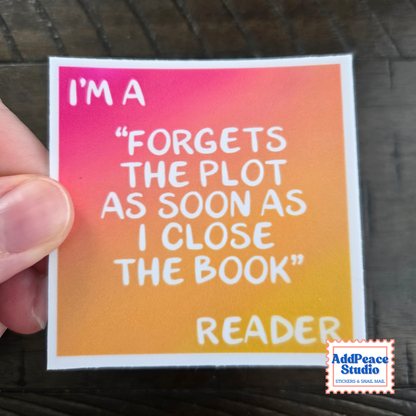 A hand holding a square sticker with a pink-to-orange gradient background and white text that reads, "I'M A 'FORGETS THE PLOT AS SOON AS I CLOSE THE BOOK' READER." A small AddPeace Studio logo appears in the bottom right corner.