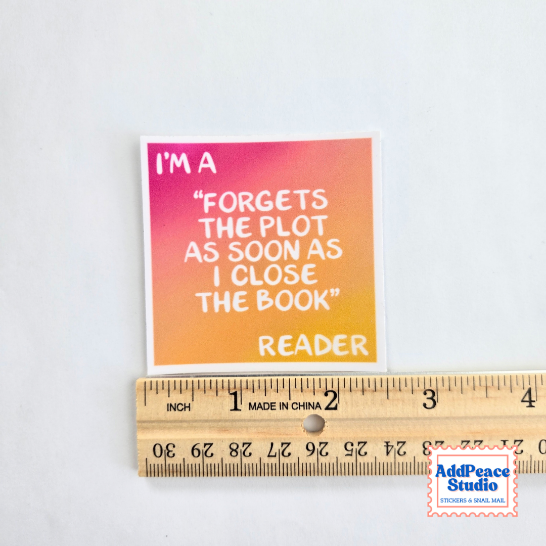 A square sticker with a pink-to-orange gradient background and white text reading, "I'M A 'FORGETS THE PLOT AS SOON AS I CLOSE THE BOOK' READER," shown above a wooden ruler for scale on a white background. AddPeace Studio logo appears in the bottom right corner.