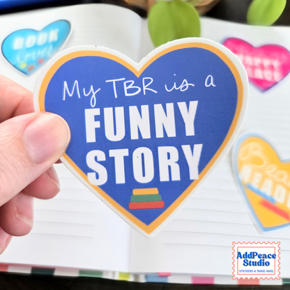 A hand holds a heart-shaped sticker that reads 'My TBR is a FUNNY STORY' in white text on a blue background with a yellow border. The sticker is shown in front of an open book with other colorful stickers visible.