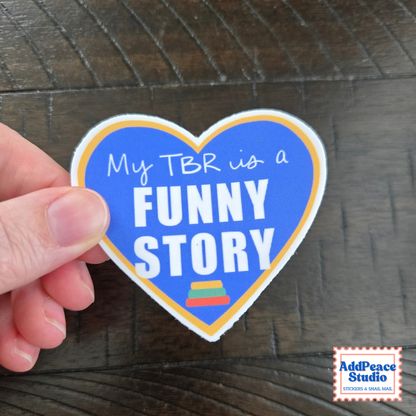 A hand holds a heart-shaped sticker with a blue background and white text that reads 'My TBR is a FUNNY STORY' against a dark wood surface. The sticker has a white border with a thin gold/yellow inner outline and a small stack of books icon at the bottom.