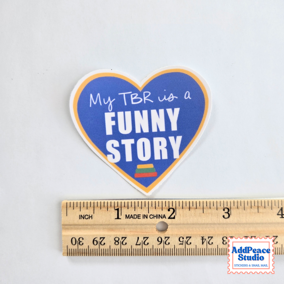 A heart-shaped sticker with the text 'My TBR is a FUNNY STORY' in white lettering on a blue background with an orange border, shown above a wooden ruler for scale on a white background.
