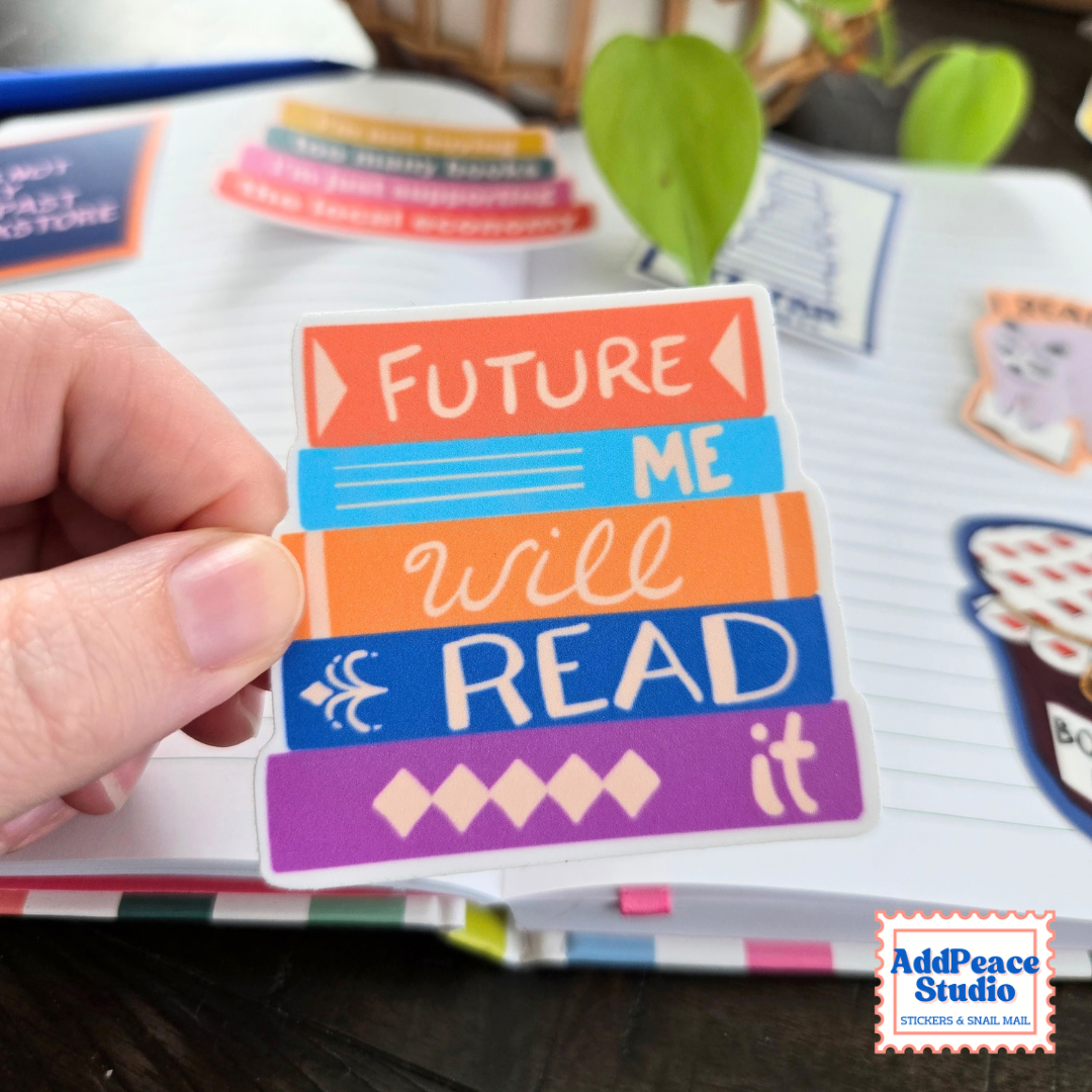 A hand holds a die-cut sticker shaped like stacked books with the words 'FUTURE ME will READ it' in colorful lettering. The sticker is shown over an open notebook, with other stickers and a plant blurred in the background.