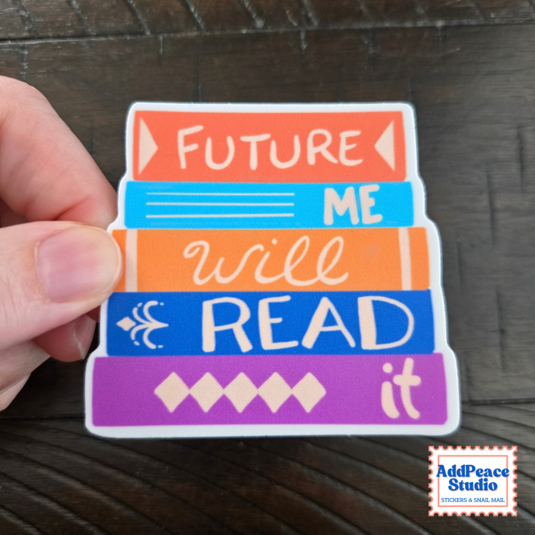 A hand holds a die-cut sticker shaped like a stack of colorful books with the words 'FUTURE ME will READ it' in white lettering, set against a dark wood background. An 'AddPeace Studio' logo is visible in the bottom right corner.