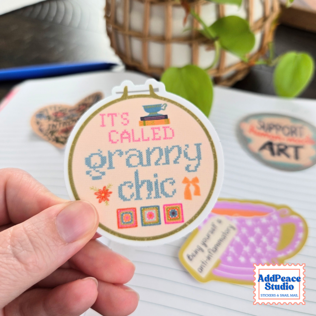 A hand holds a round embroidered-style sticker that reads “IT’S CALLED granny chic” with small decorative graphics. Other stickers are visible on a notebook page in the background, along with a plant and a woven basket.