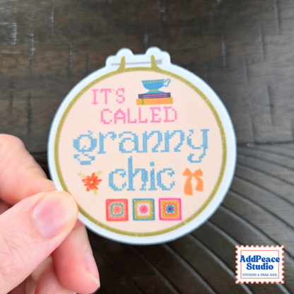 A hand holding a round sticker that reads 'IT'S CALLED granny chic' in pink and blue lettering, with small decorative illustrations, against a dark wood background. AddPeace Studio logo is visible in the bottom right corner.
