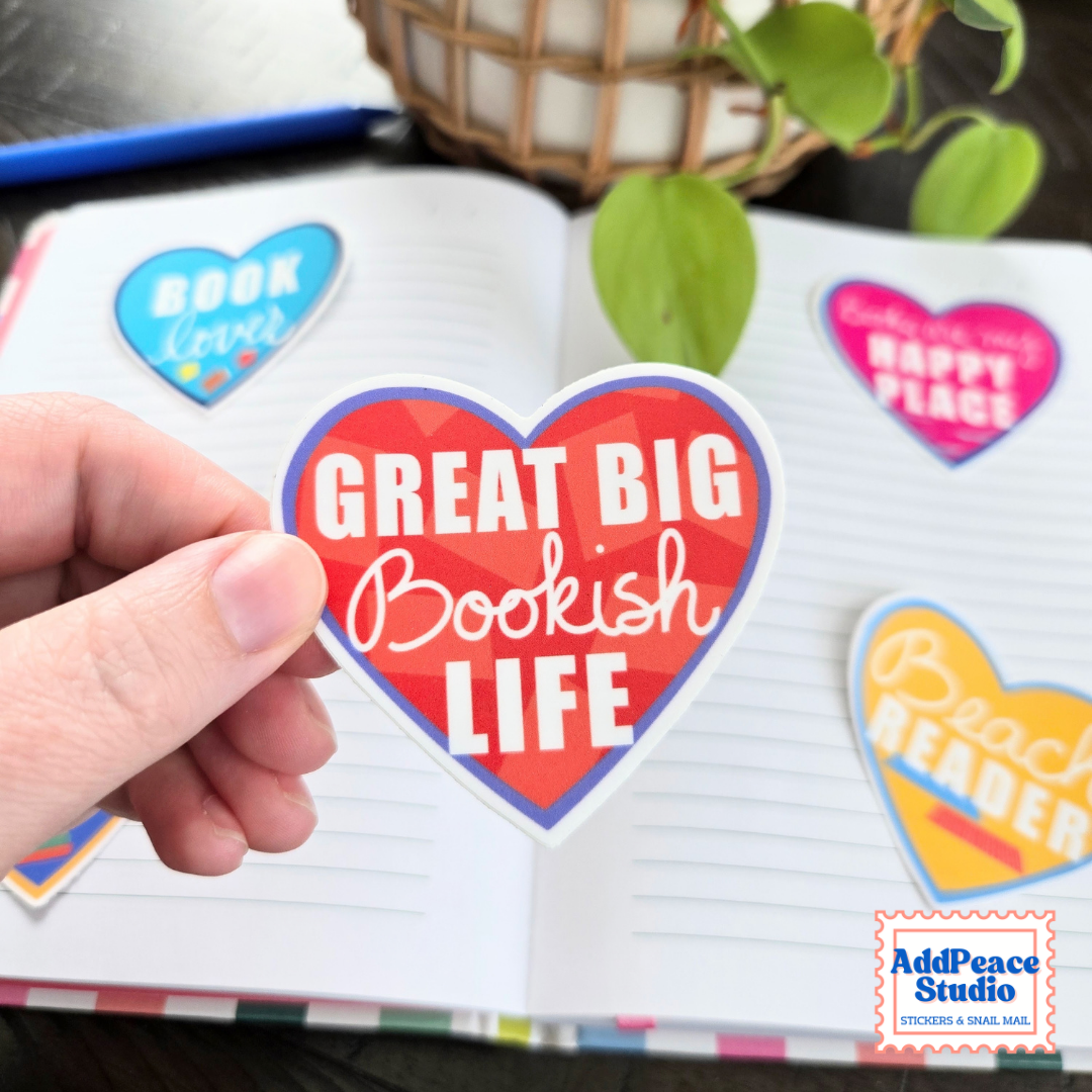 A hand holds a heart-shaped sticker that reads 'GREAT BIG Bookish LIFE' in front of an open notebook with other colorful heart stickers on the pages. The AddPeace Studio logo is visible in the bottom right corner.
