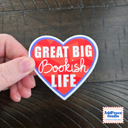 A hand holds a red heart sticker with white text saying Great big bookish life over a dark background with the AddPeace Studio logo in the bottom right corner