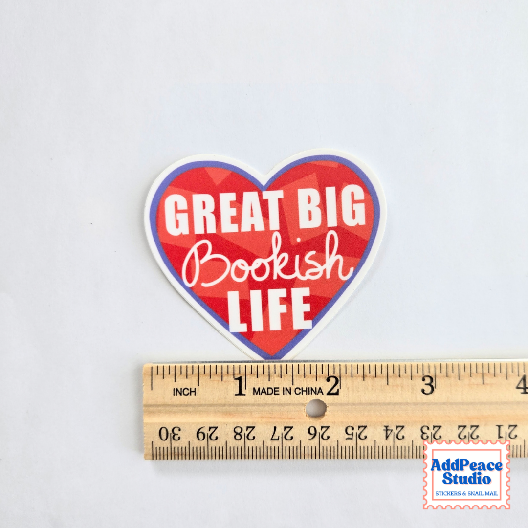 A heart-shaped sticker with the text 'GREAT BIG Bookish LIFE' in white lettering over a red and blue geometric pattern, shown above a wooden ruler to indicate size. A 'AddPeace Studio' watermark is visible in the bottom right corner.