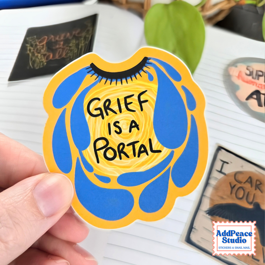 A hand holds a die-cut sticker featuring a stylized eye crying blue tears and the text 'GRIEF IS A PORTAL' in black. The background shows a white desk with other stickers, a green leaf, and a notebook.