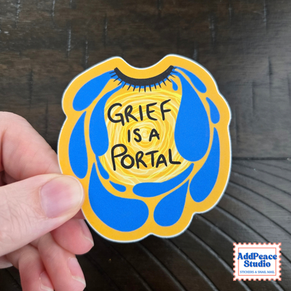 A hand holds a die-cut sticker featuring a yellow and blue abstract design with the text 'GRIEF IS A PORTAL' in black. The background is a dark wood surface, and a 'AddPeace Studio' logo is visible in the bottom right corner.