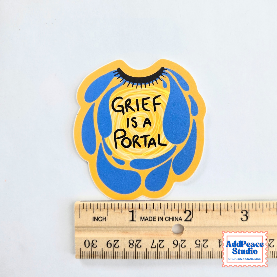 A die-cut sticker with the text 'GRIEF IS A PORTAL' in black over a yellow and blue abstract design, shown above a wooden ruler for scale on a white background.
