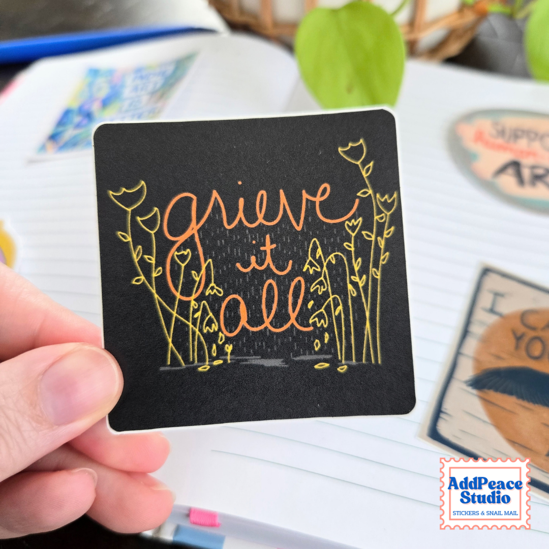 A hand holds a square black sticker with the phrase 'grieve it all' in copper-orange script and yellow floral line art. The background shows a notebook and other stickers, with a visible AddPeaceStudio logo in the bottom right corner.