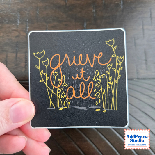 A hand holds a square black sticker featuring the phrase 'grieve it all' in orange script and yellow floral line art, set against a dark wooden background. An 'AddPeace Studio' logo is visible in the bottom right corner.