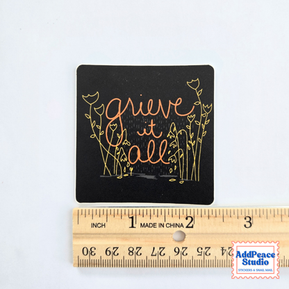 A square black sticker with the phrase 'grieve it all' in cursive and gold floral illustrations, shown above a wooden ruler for scale on a white background.