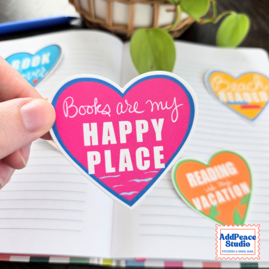 A hand holds a heart-shaped sticker that reads 'Books are my HAPPY PLACE' in white lettering on a bright pink background with a blue border. Other colorful heart stickers are placed on an open notebook, with a plant and basket in the background.