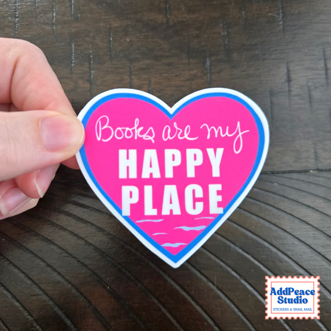 A hand holds a heart-shaped sticker that reads 'Books are my HAPPY PLACE' in white text on a bright pink background with a blue border, set against a dark wood surface. AddPeace Studio logo is visible in the bottom right corner.