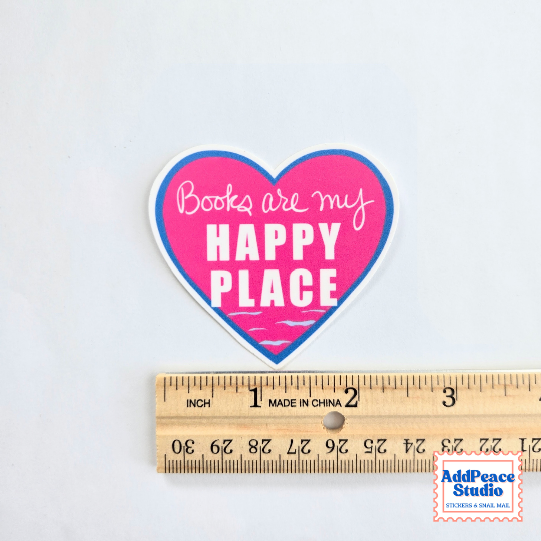 A heart-shaped sticker with the text 'Books are my HAPPY PLACE' in white lettering, featuring a pink center, blue border, and white outer outline. The sticker is displayed above a wooden ruler on a plain white background to show its size.