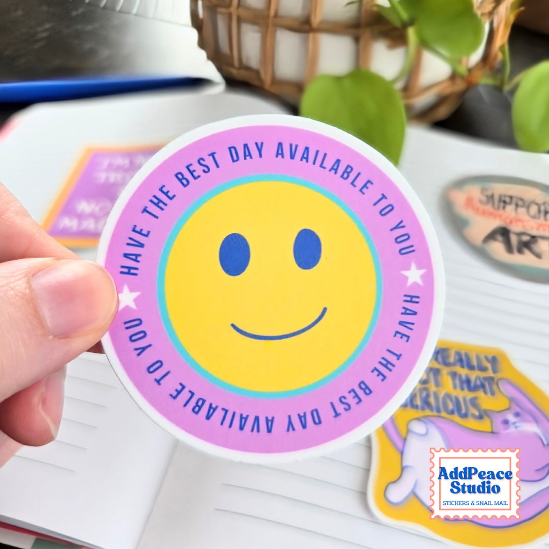A hand holds a round sticker featuring a yellow smiley face surrounded by a pink and purple border with the text 'HAVE THE BEST DAY AVAILABLE TO YOU'. The background shows a notebook and other stickers, with a plant in the soft-focus background.