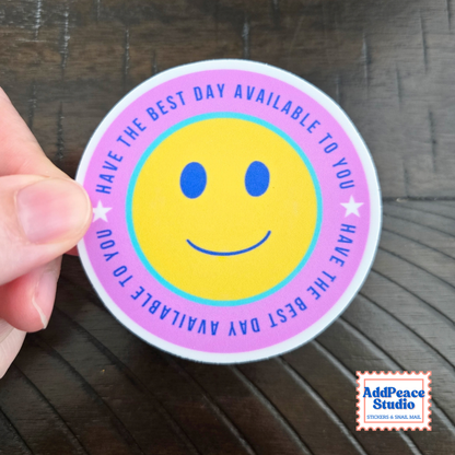 A hand holding a round sticker with a yellow smiley face and the text 'HAVE THE BEST DAY AVAILABLE TO YOU' in blue on a pink and light blue border, set against a dark wood background.