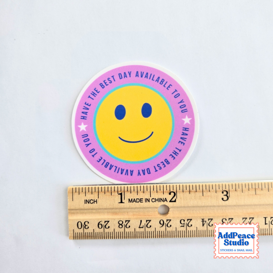 A round sticker with a yellow smiley face and a pink outer ring reading 'HAVE THE BEST DAY AVAILABLE TO YOU' is shown above a wooden ruler for scale on a white background.