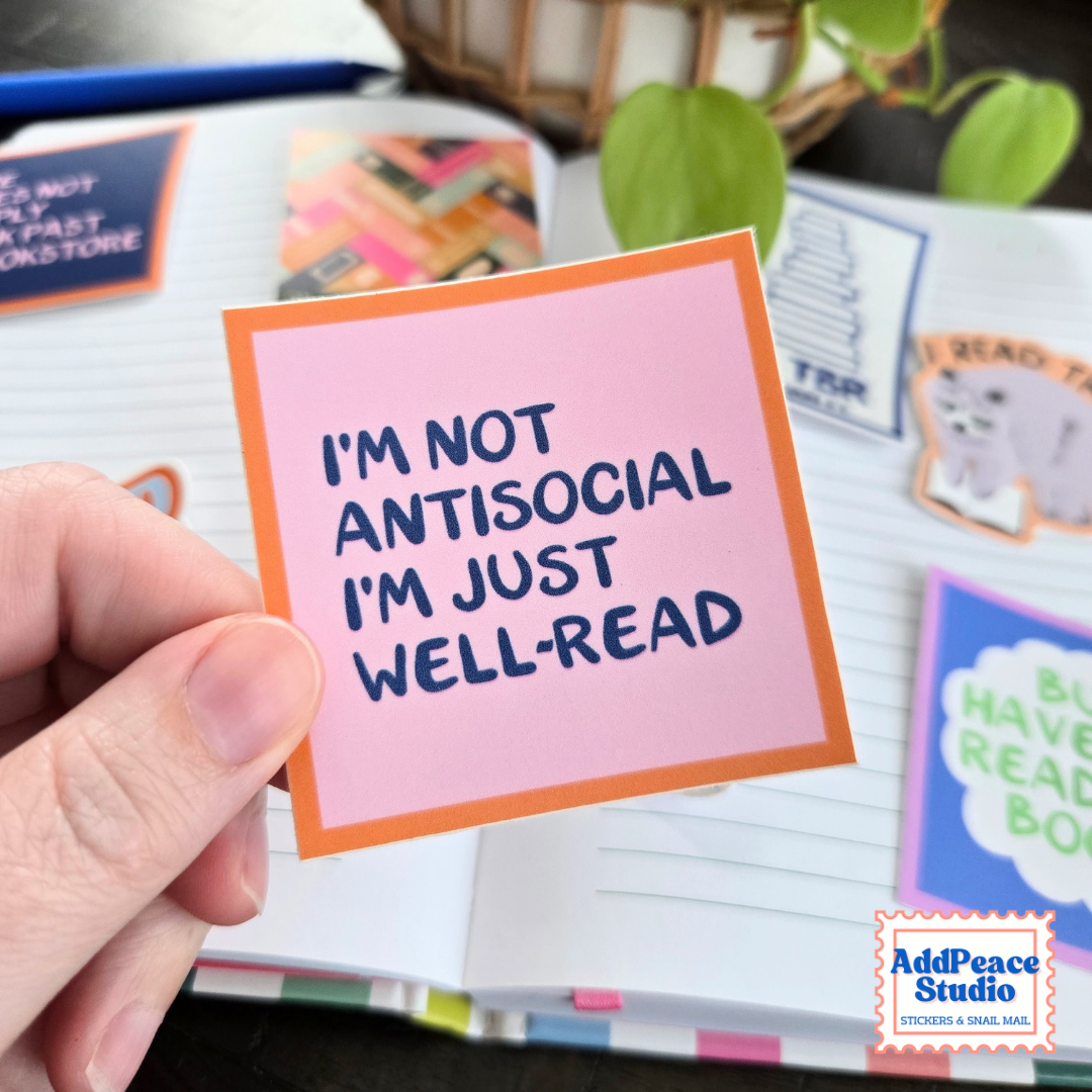 A hand holds a pink sticker with an orange border that reads, 'I'M NOT ANTISOCIAL I'M JUST WELL-READ' in dark blue text. The background shows an open notebook with colorful stickers and books, and a plant leaf is visible.