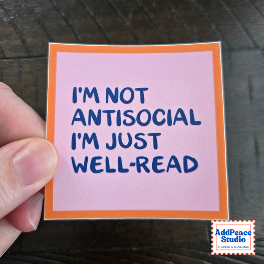 A hand holds a square sticker with a pink background, orange border, and navy blue text that reads 'I'M NOT ANTISOCIAL I'M JUST WELL-READ'. A small 'AddPeace Studio' brand mark appears in the bottom right corner.