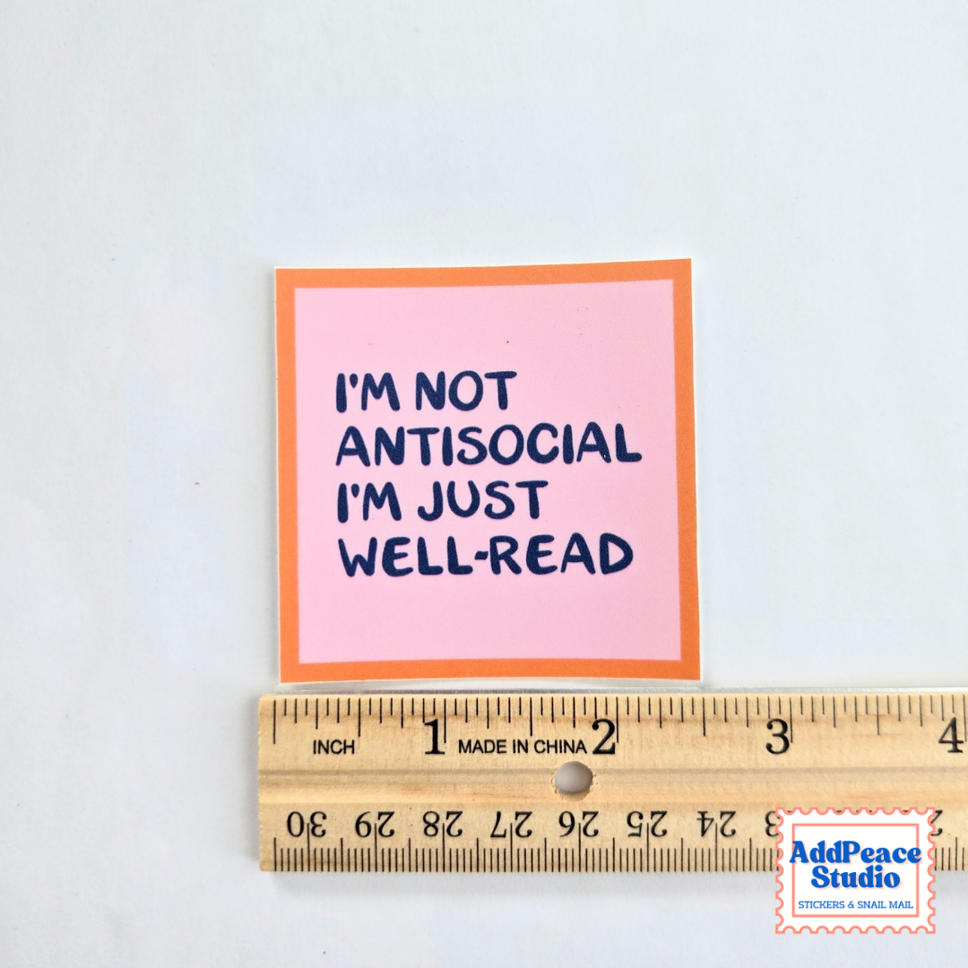 A square pink sticker with an orange border and navy blue text reading 'I'M NOT ANTISOCIAL I'M JUST WELL-READ' is shown above a wooden ruler on a white background. An 'AddPeace Studio' logo is in the bottom right corner.