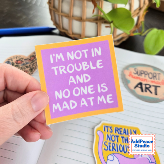 A hand holds a square sticker with a purple background and yellow border, featuring the text 'I'M NOT IN TROUBLE AND NO ONE IS MAD AT ME' in white. The scene is a styled desk setup with other stickers, a notebook, and a plant in the background.
