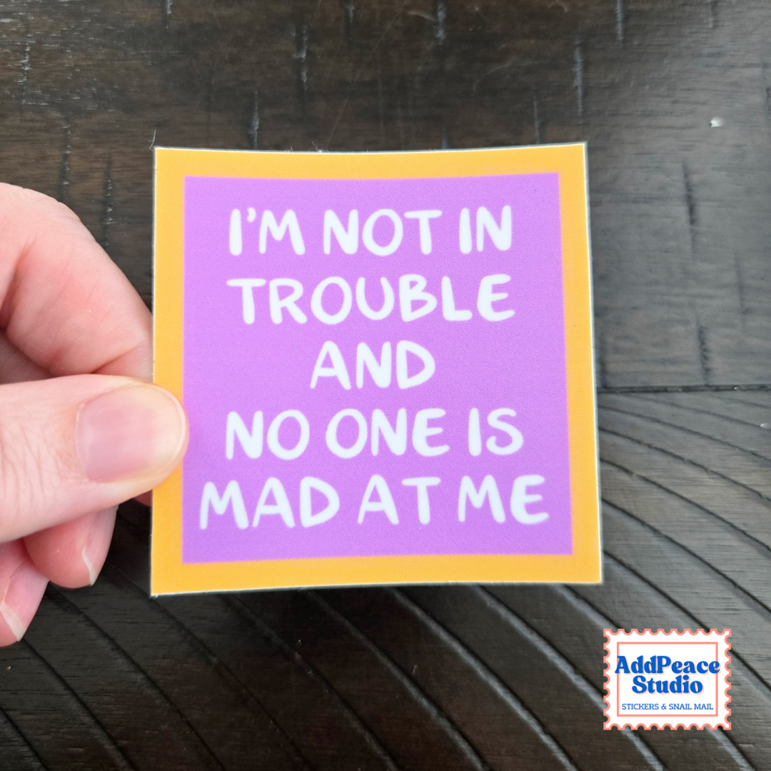A hand holds a square sticker with a purple background, yellow border, and white text that reads, 'I'M NOT IN TROUBLE AND NO ONE IS MAD AT ME' on a dark wood surface. AddPeace Studio logo is visible in the bottom right corner.