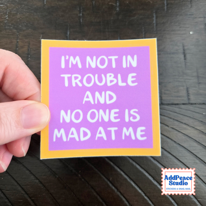 A hand holds a square sticker with a purple background, yellow border, and white text that reads, 'I'M NOT IN TROUBLE AND NO ONE IS MAD AT ME' on a dark wood surface. AddPeace Studio logo is visible in the bottom right corner.