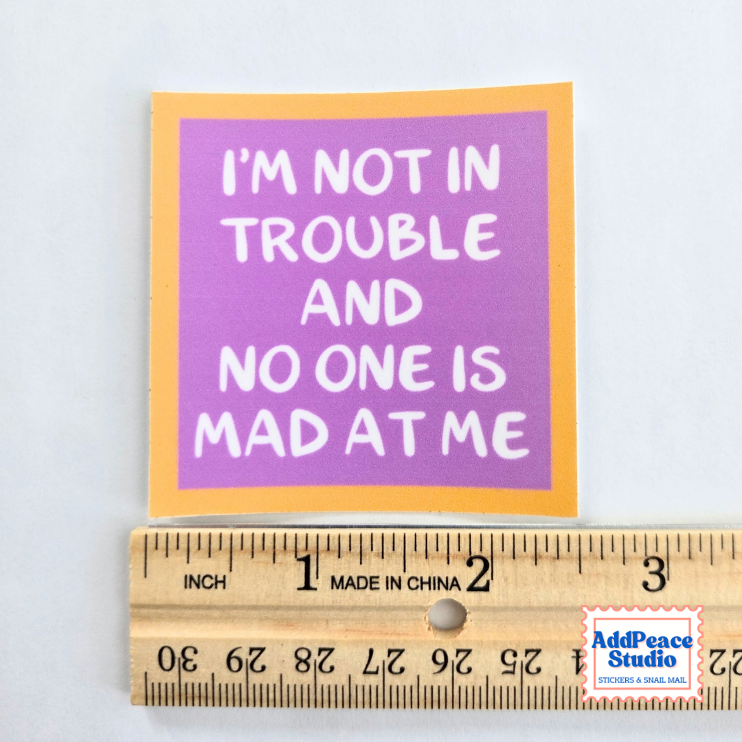 A square sticker with the text 'I'M NOT IN TROUBLE AND NO ONE IS MAD AT ME' in white lettering on a purple background with a yellow border, shown next to a wooden ruler for scale on a white background.