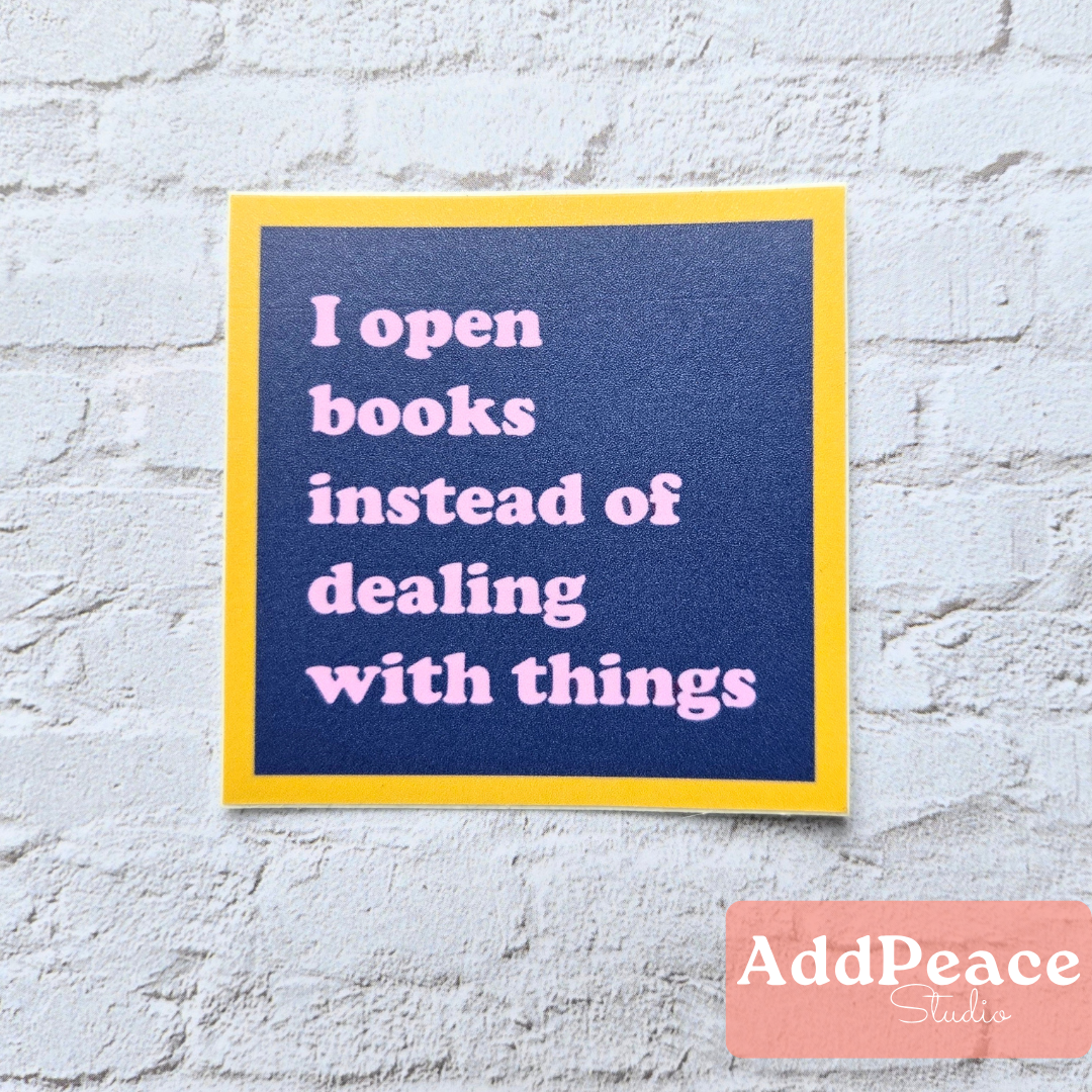 Sticker with text 'I open books instead of dealing with things' on a blue background with yellow border, against a light gray brick wall. Brand 'AddPeace Studio' visible.