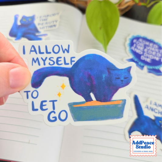 A hand holds a die-cut sticker featuring a blue cat standing in a litterbox with the text 'I ALLOW MYSELF TO LET GO' in blue lettering. The sticker is shown over an open notebook, with other matching stickers visible in the background.
