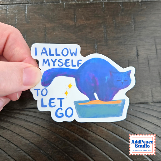 A hand holds a die-cut sticker featuring a blue cat standing on a green litter box with the text 'I ALLOW MYSELF TO LET GO' on a dark wood background. An 'AddPeace Studio' logo is visible in the bottom right corner.