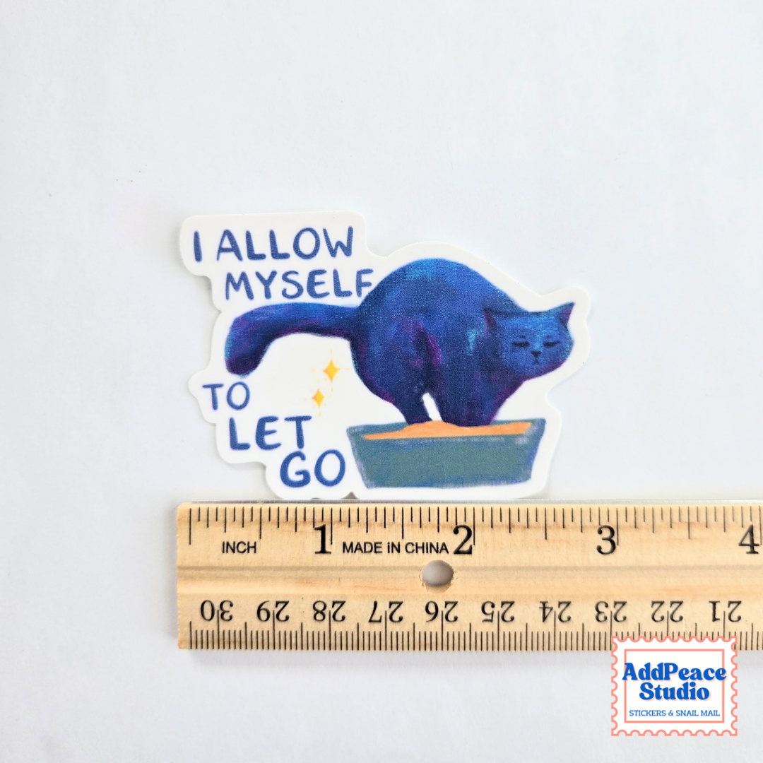 A die-cut sticker featuring a blue cat standing in a litterbox with the text 'I ALLOW MYSELF TO LET GO' in blue lettering, shown above a wooden ruler for scale on a white background.