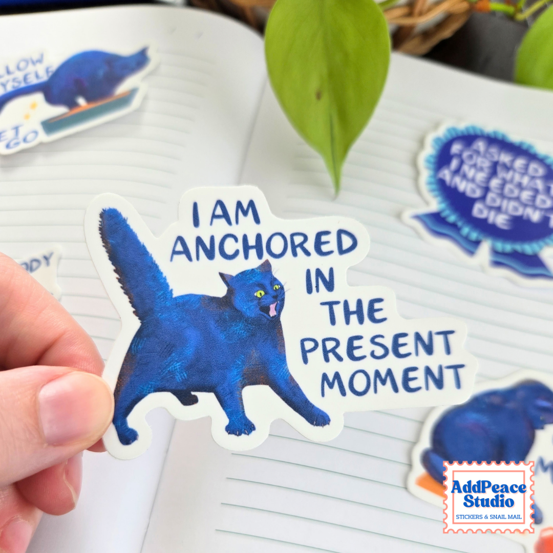 A hand holds a die-cut sticker featuring a blue cat and the text 'I AM ANCHORED IN THE PRESENT MOMENT' over an open notebook with other stickers visible in the background.