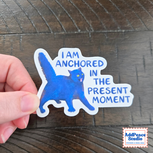 A hand holds a die-cut sticker featuring a blue cat and the text 'I AM ANCHORED IN THE PRESENT MOMENT' on a dark wood background. The AddPeace Studio logo is visible in the bottom right corner.