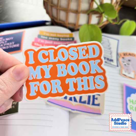 A hand holds a die-cut sticker that reads 'I CLOSED MY BOOK FOR THIS' in light blue text on an orange background. The sticker is shown in front of an open notebook with other colorful stickers scattered around, and a plant and basket are blurred in the background.