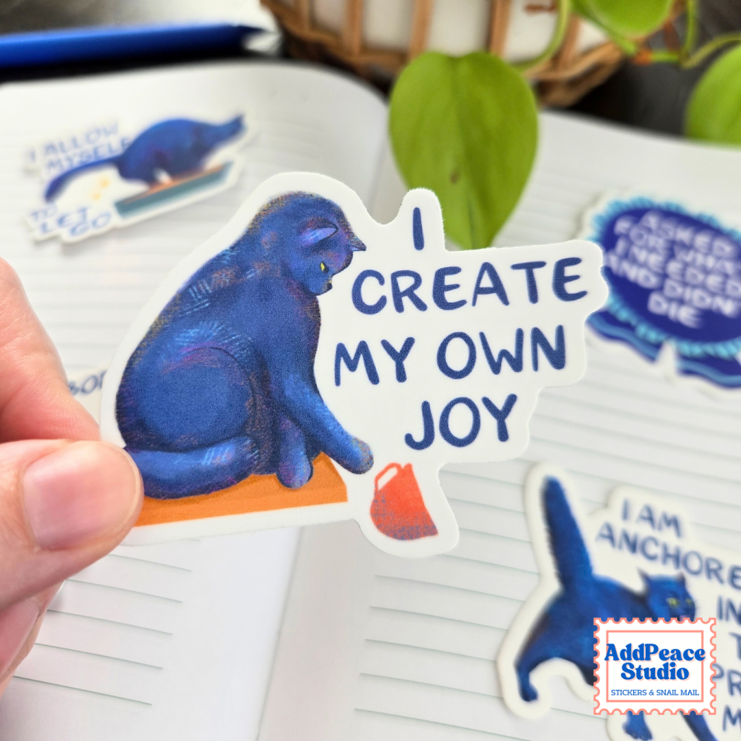 A hand holds a die-cut sticker featuring a blue cat sitting on an orange surface next to the text 'I CREATE MY OWN JOY' in blue lettering on a white background. Other matching stickers are visible on lined paper in the background, with a plant and basket blurred in the distance.