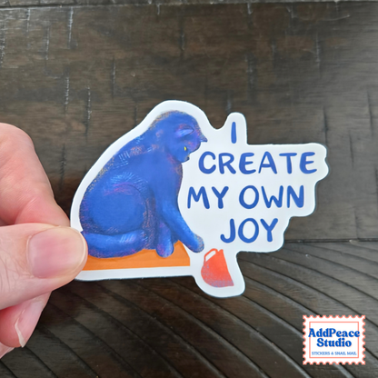 A hand holds a white die-cut sticker featuring a blue cat sitting on an orange block with the text 'I CREATE MY OWN JOY' in blue lettering. The sticker is photographed over a dark brown wooden surface, with an 'AddPeace Studio' logo in the bottom right corner.
